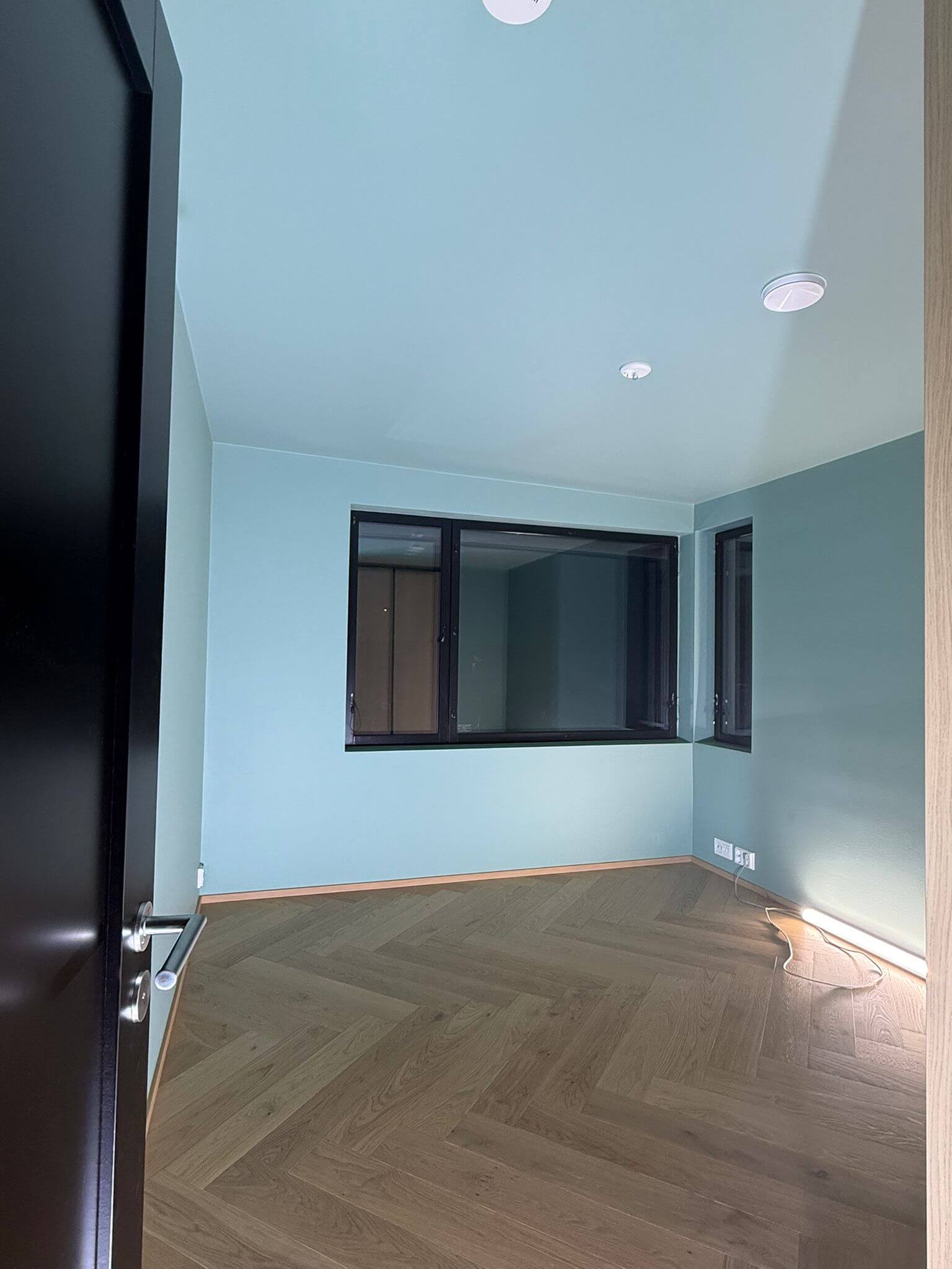 Huoneessa on kalanruotokuvioinen parketti, turkoosit seinät, mustat ikkunanpuitteet ja musta ovi.