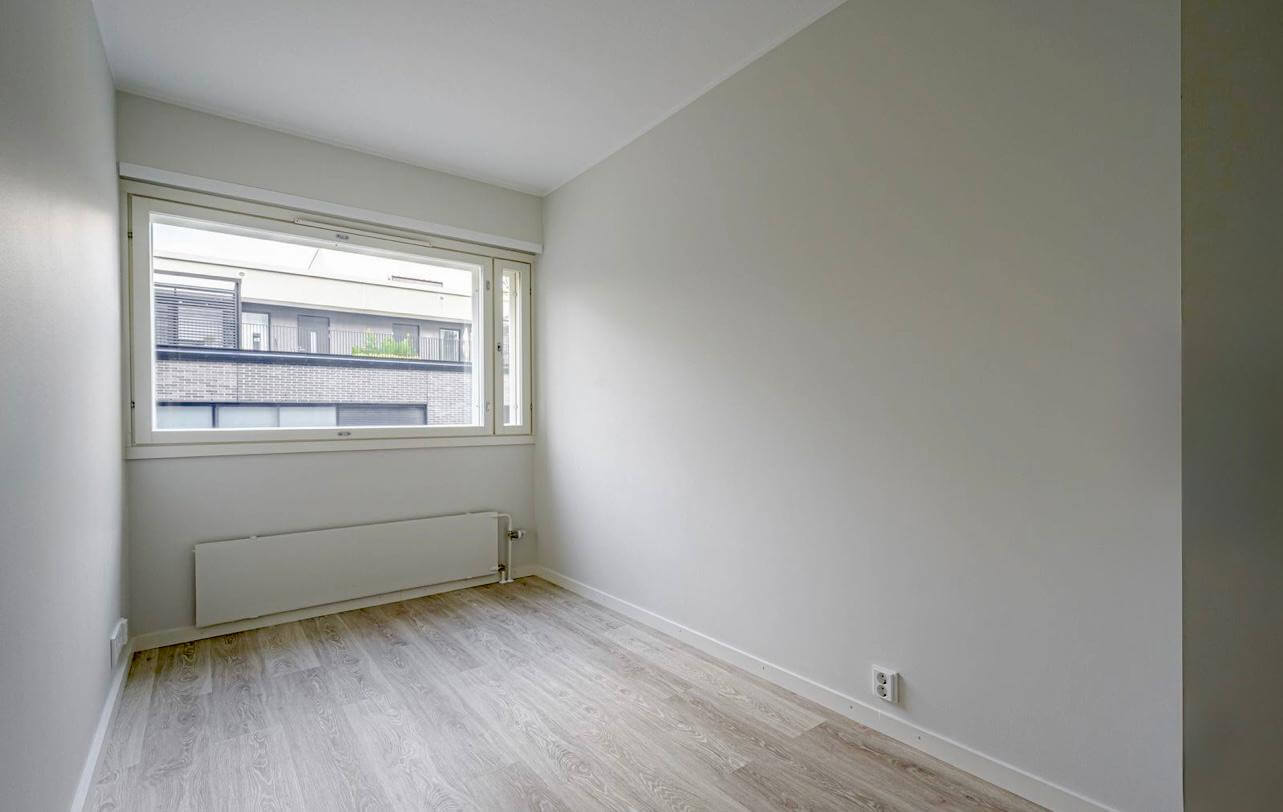 Vaalea huone, jossa on ikkuna, patteri ja laminaattilattia. Seinät ovat vaaleanharmaat ja valaistus on luonnollista.