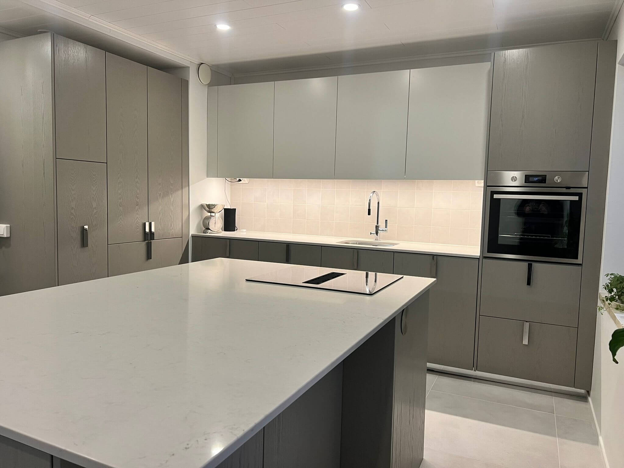 Moderni keittiö, jossa on harmaat kaapit, valkoinen saareke ja ruostumattomasta teräksestä valmistettu uuni. Työtasot ovat vaaleat ja valaistus on kirkas.