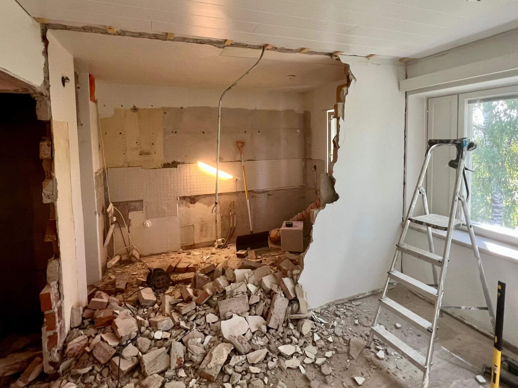 Remonttityömaa, jossa on tiiliä lattialla, seinät osittain purettu ja tikkaat ikkunan vieressä.