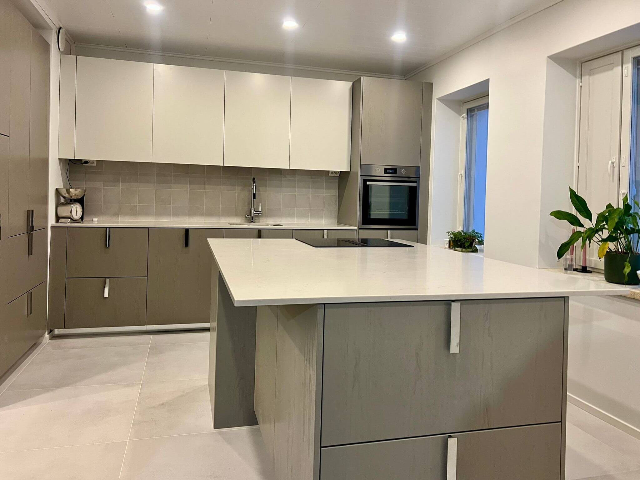 Moderni keittiö, jossa on harmaat alakaapit, valkoiset yläkaapit, valkoinen saareke ja kasveja ikkunalaudalla.