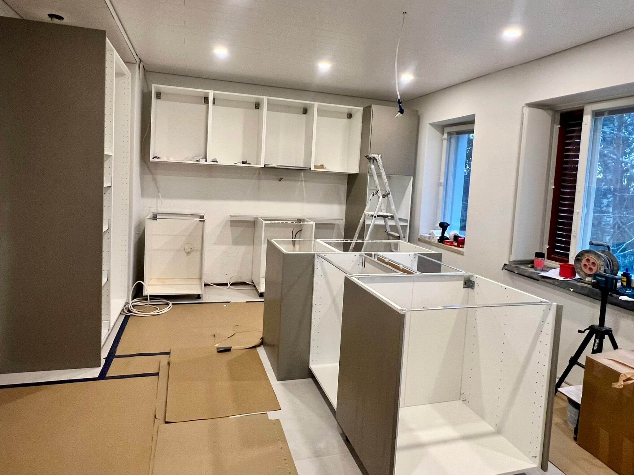 Keskeneräinen keittiö asennus, jossa on harmaita ja valkoisia kaappeja, pahvisuoja lattialla ja valaistus katossa.