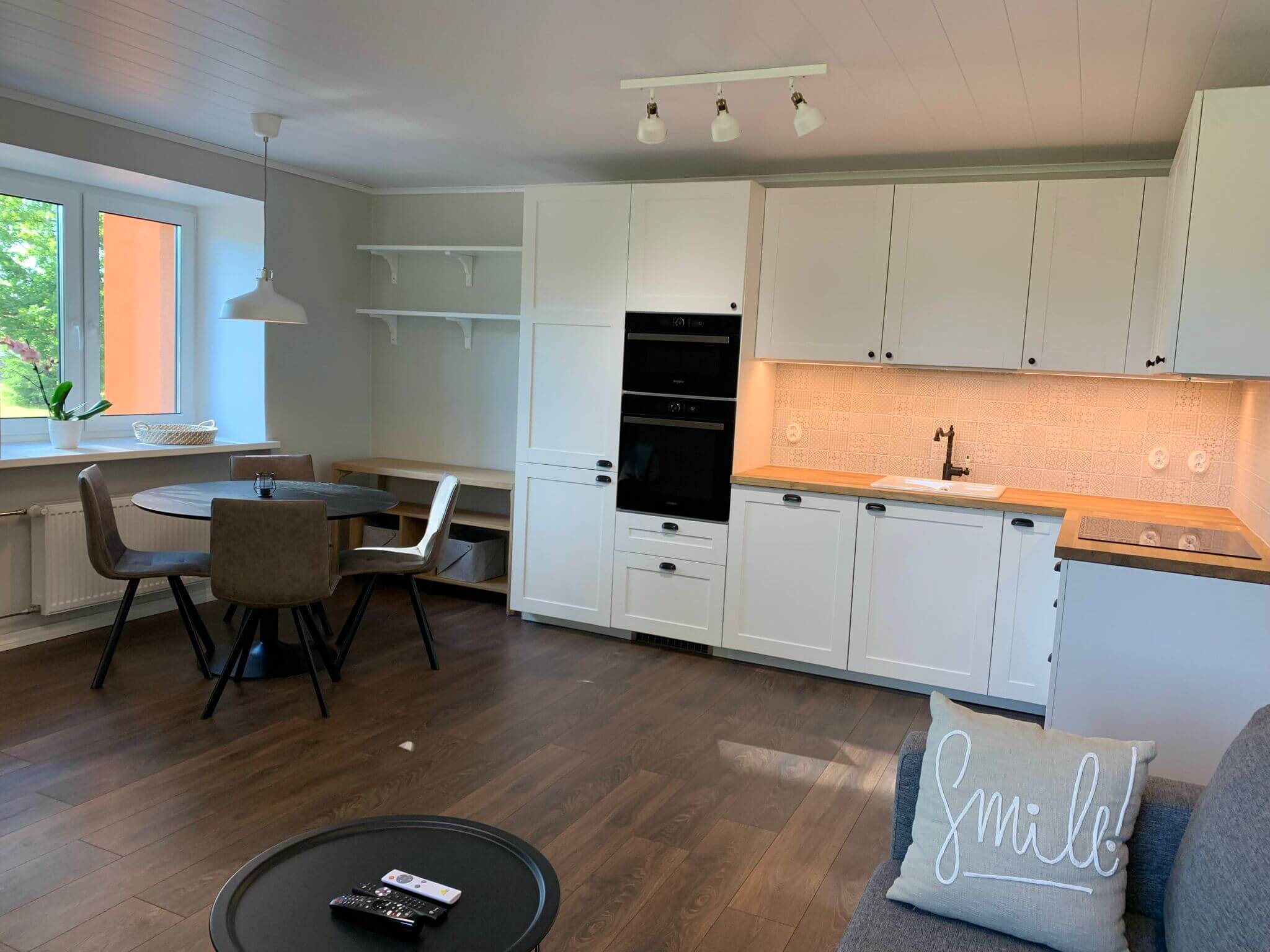 Sisätiloissa on keittiö, jossa on valkoiset kaapit, puinen työtaso, ruokapöytä ja tuolit sekä sohva, jossa on "Smile!"-tyyny.