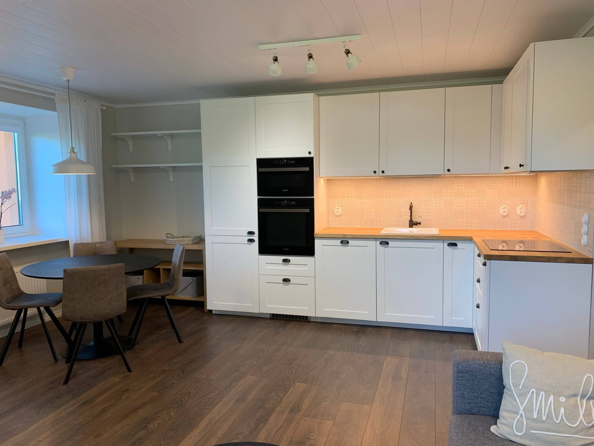 Keittiönäkymä, jossa on valkoiset kaapit, puulattia, pyöreä pöytä ja harmaat tuolit. Valaisimet katossa ja pöydän yläpuolella.