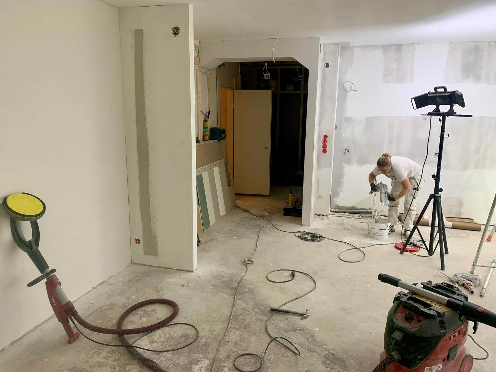 Huone remontissa, jossa työntekijä sekoittaa maalia, lattialla työkaluja ja valaisin valaisee tilaa.