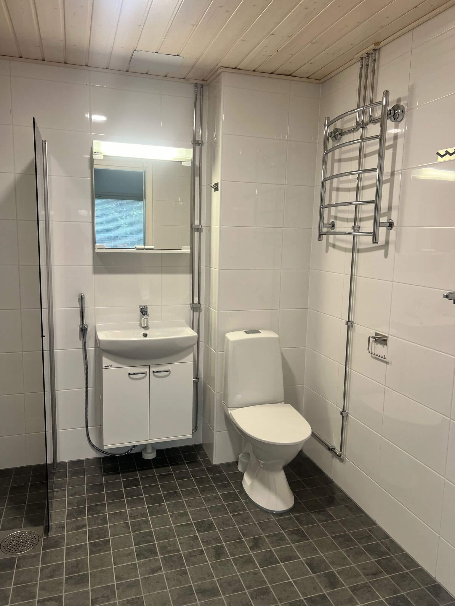 Kylpyhuoneessa on valkoinen pesuallas, peili, wc-istuin, pyyhekuivain ja harmaa laattalattia.