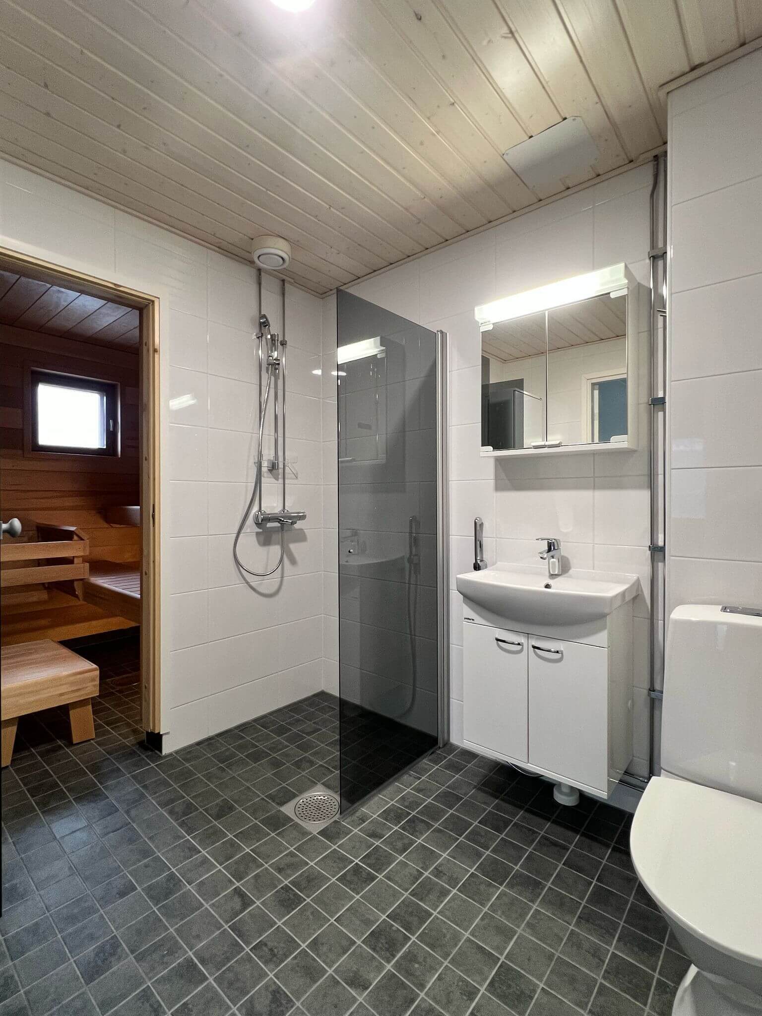 Kylpyhuoneessa on sauna, suihku, pesuallas ja wc. Harmaat lattialaatat ja valkoiset seinälaatat. Puupaneloitu katto ja kirkas valaistus.