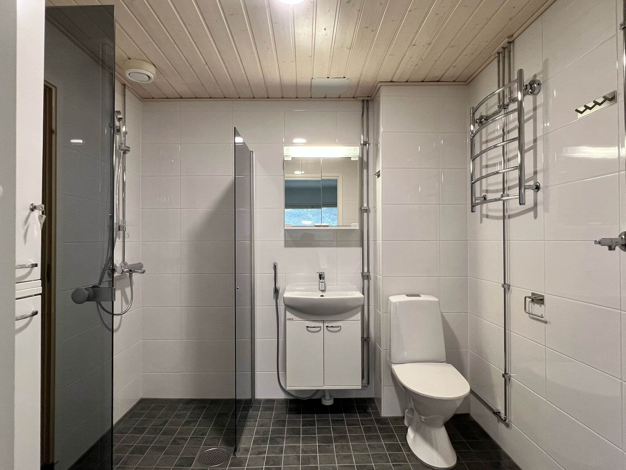 Kylpyhuoneessa on harmaat lattialaatat, valkoiset seinälaatat, puinen katto, suihkukaappi, peili, valkoinen pesuallas ja wc-istuin.