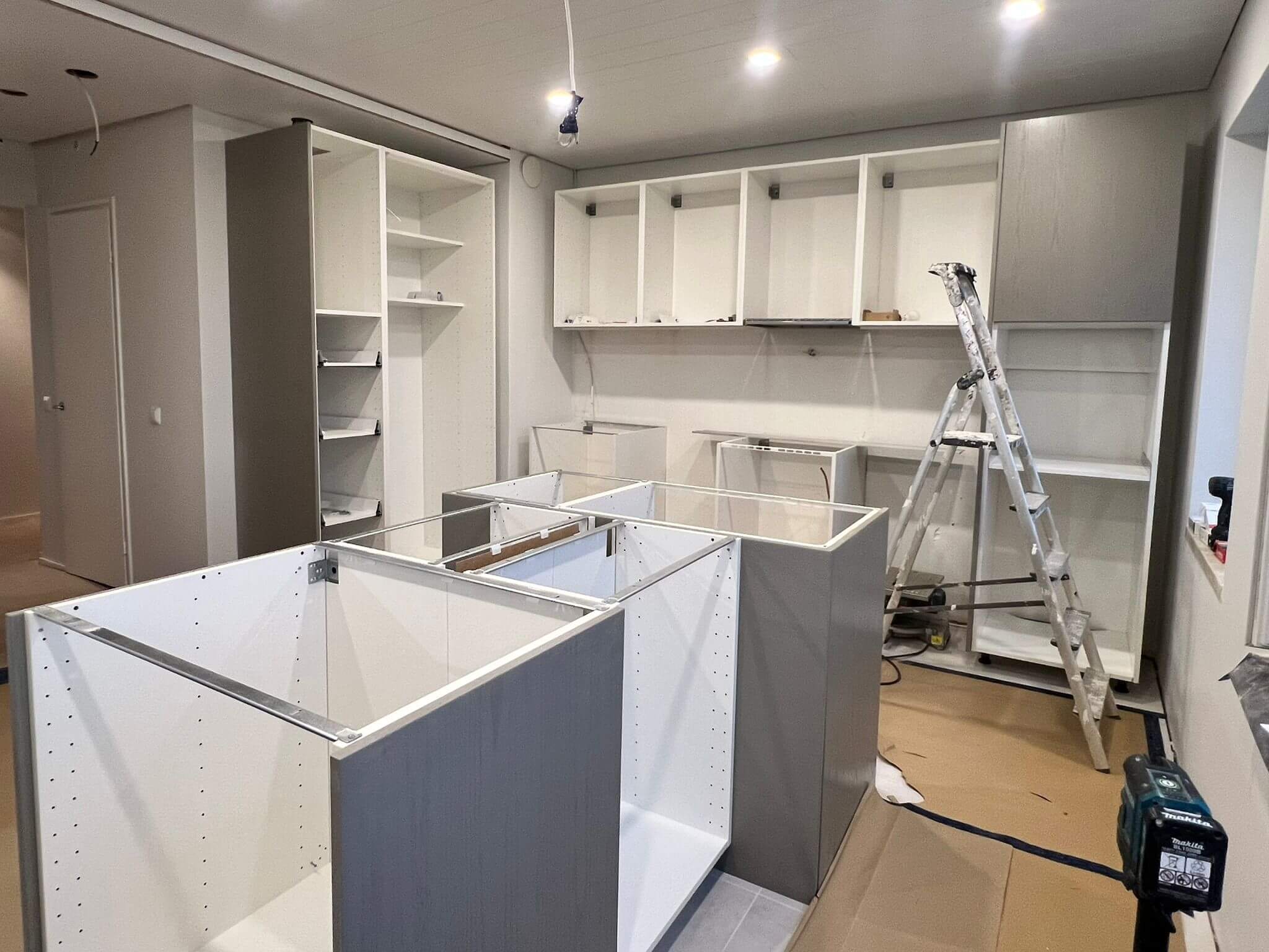 Keskeneräinen keittiö asennuksessa, harmaat kaapit, valkoinen sisustus, valaistus katossa.