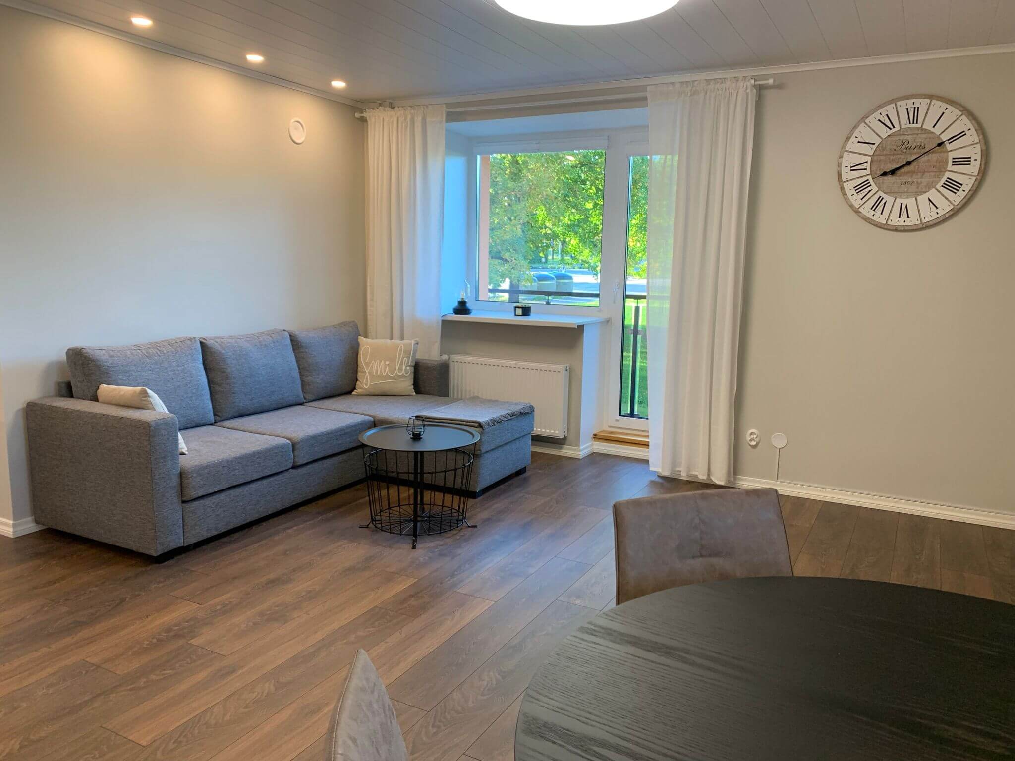 Olohuone, jossa on harmaa kulmasohva, pyöreä sohvapöytä, suuri seinäkello ja tumma puulattia.