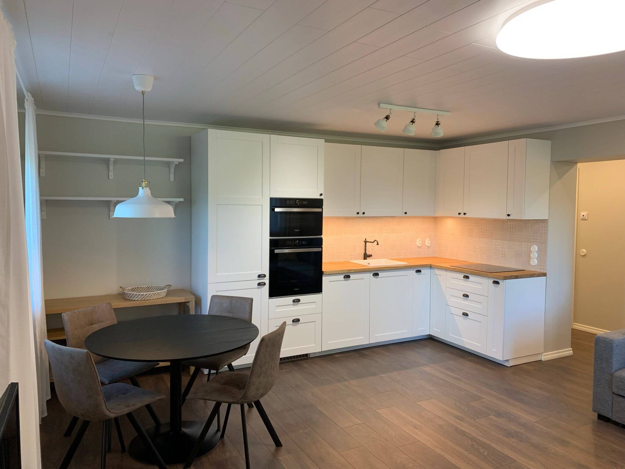 Vaaleassa huoneessa on valkoiset keittiökaapit, tumma pyöreä pöytä ja neljä harmaata tuolia, sekä puulattia ja valkoinen kattovalaisin.