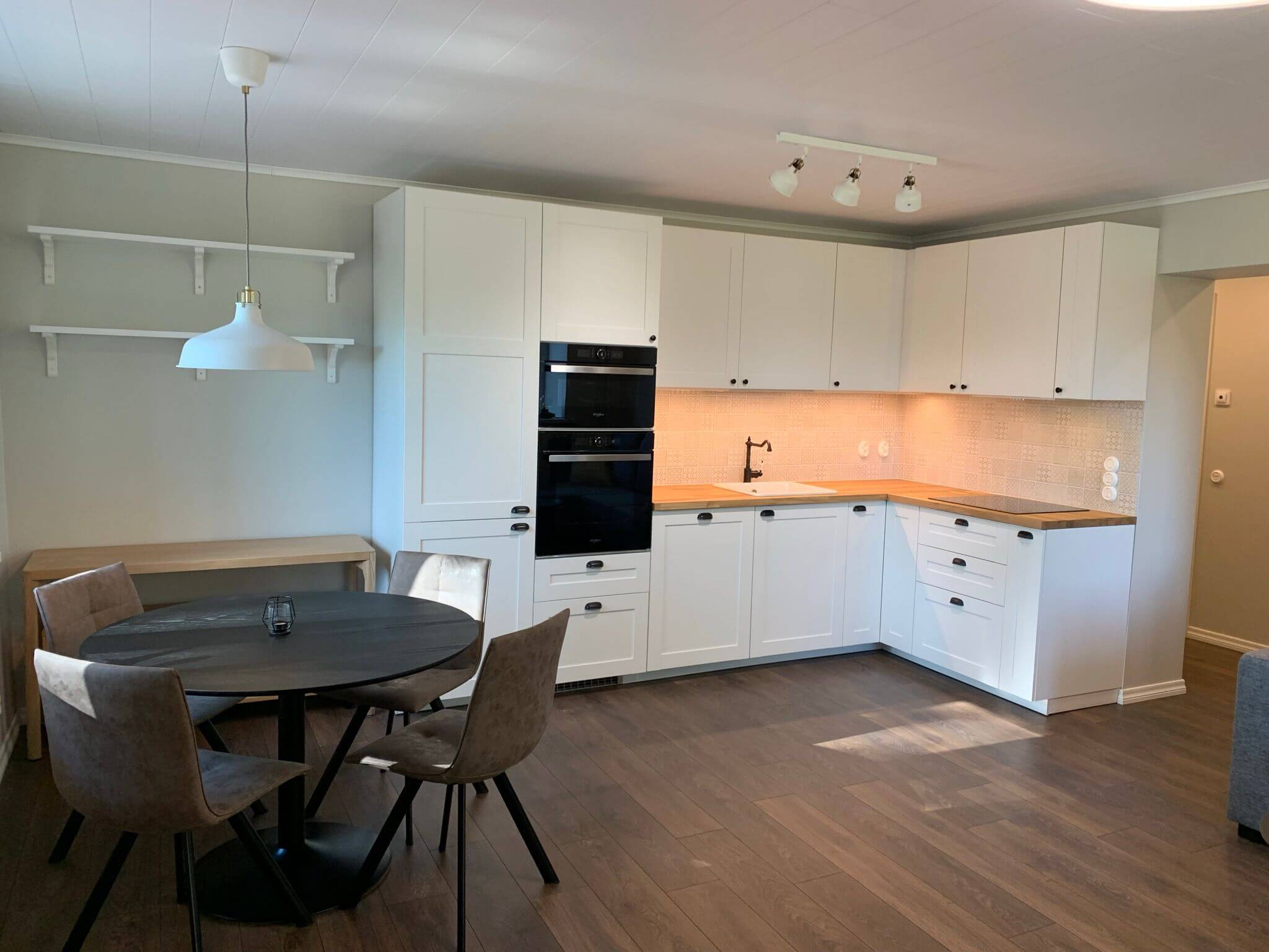 Keittiönäkymä, jossa on valkoiset kaapit, tumma ruokapöytä ja neljä tuolia, sekä valkoinen riippuvalaisin.