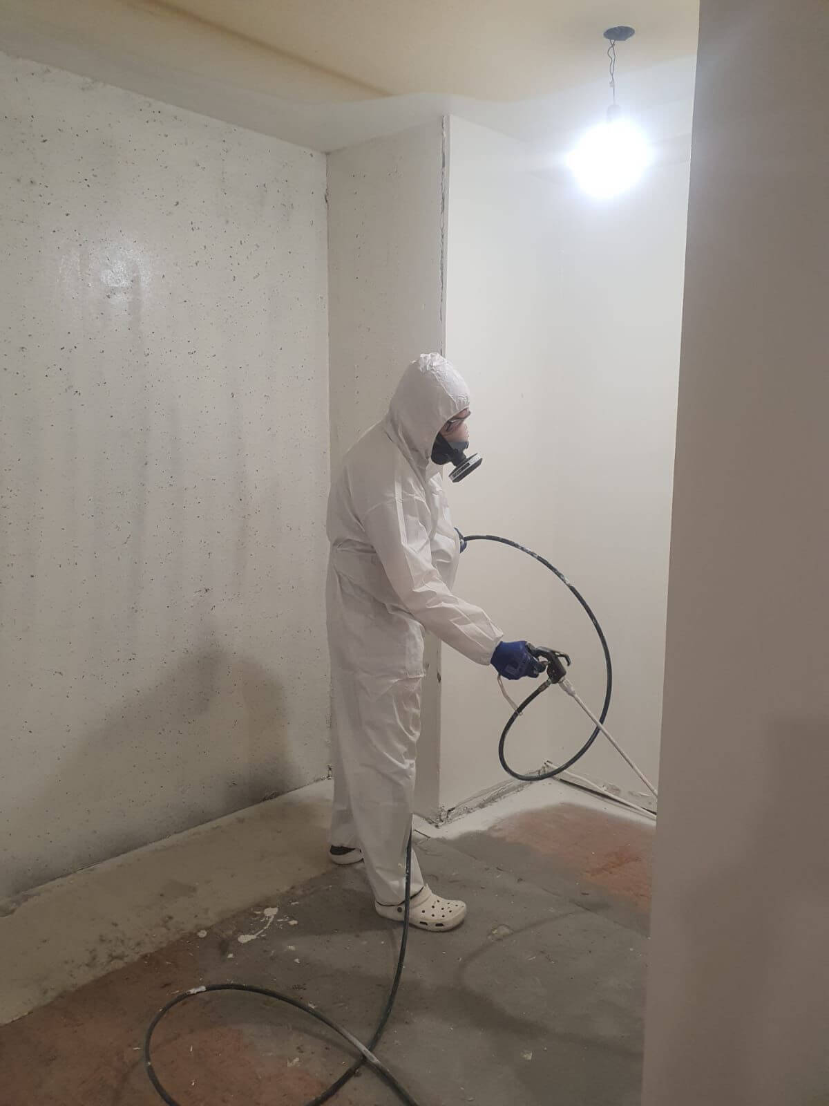 Henkilö suojapuvussa ja hengityssuojaimessa ruiskuttaa maalia huoneessa, jossa on valkoiset seinät ja betonilattia. Huoneessa on kirkas valo katossa.