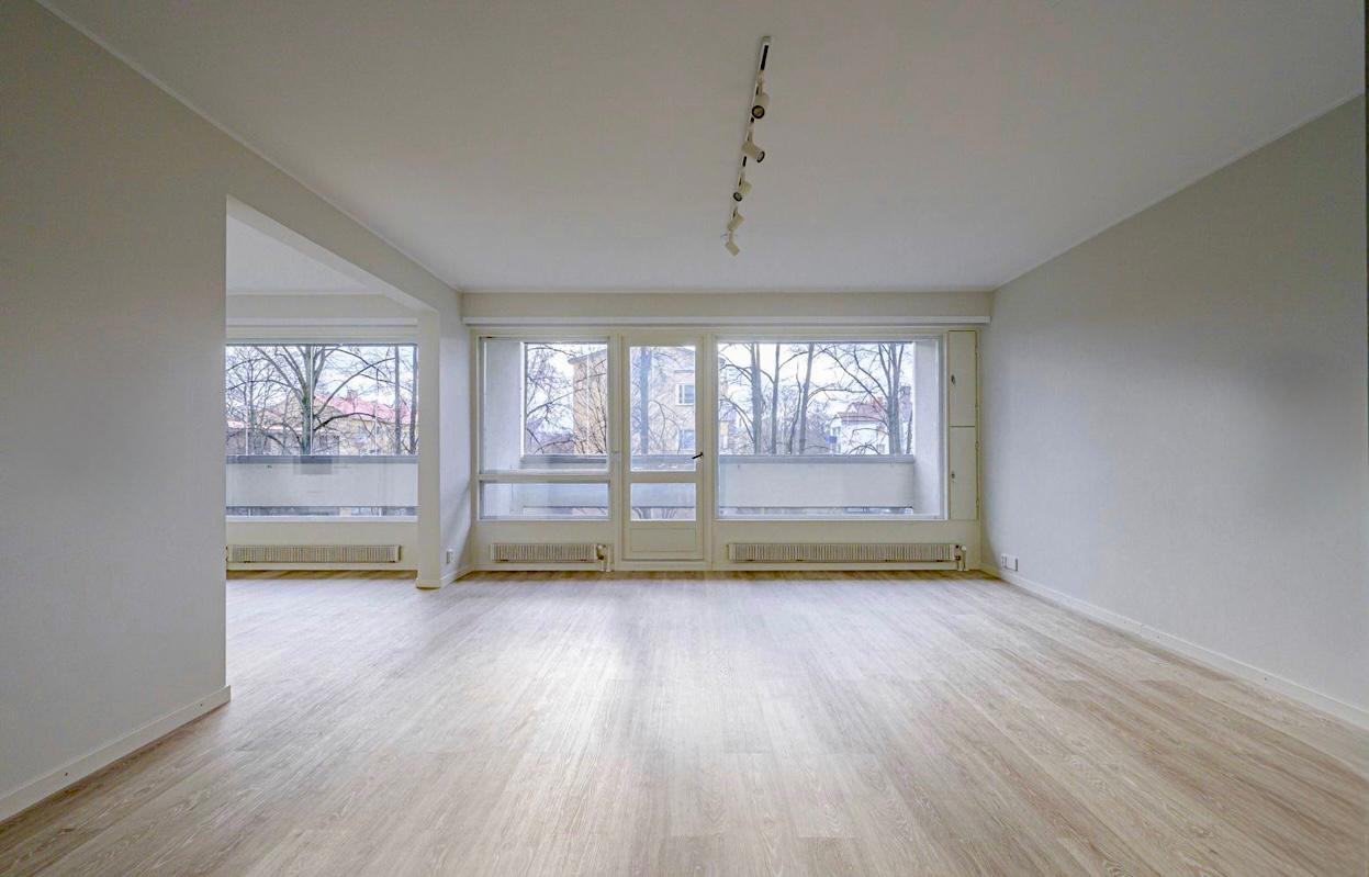 Tyhjä, valoisa huone, jossa on suuret ikkunat ja vaalea puulattia. Kattovalaisin roikkuu katosta.