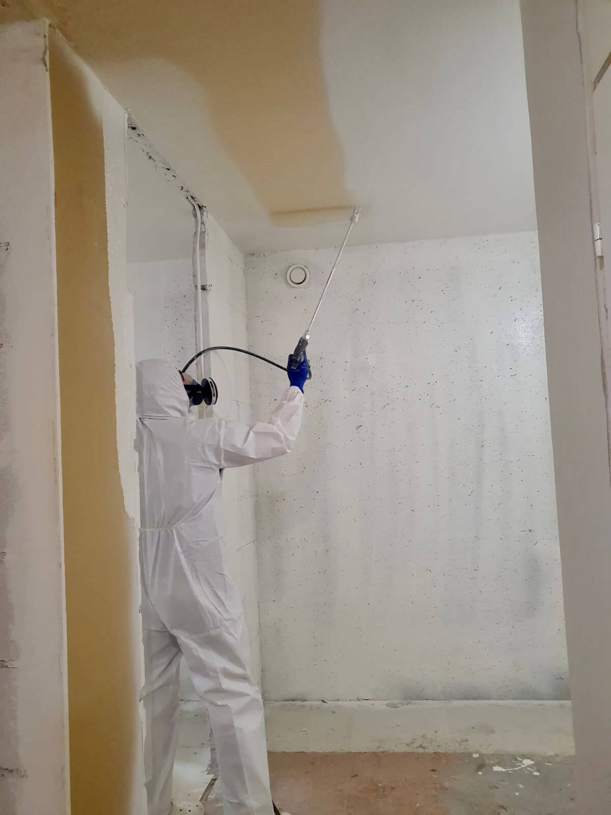 Henkilö suojapuvussa ruiskuttaa kattoa, jossa on osittain maalattu beigeä maalia. Seinät ovat valkoiset ja täynnä pieniä pilkkuja.