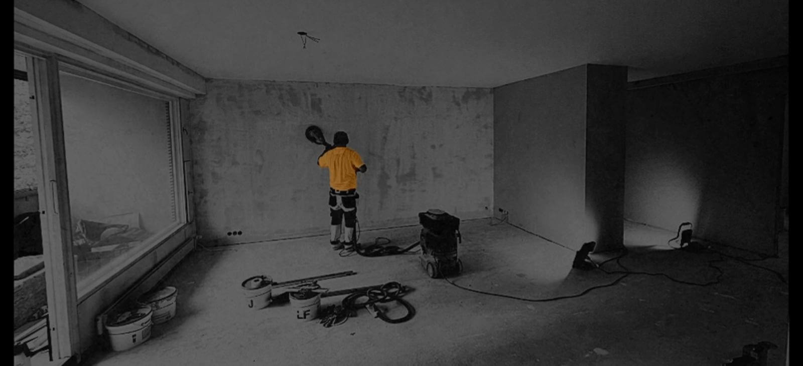 Remonttimies hioo seinää keltaisessa paidassa, työkaluja ja valaisimia lattialla, harmaa huone.