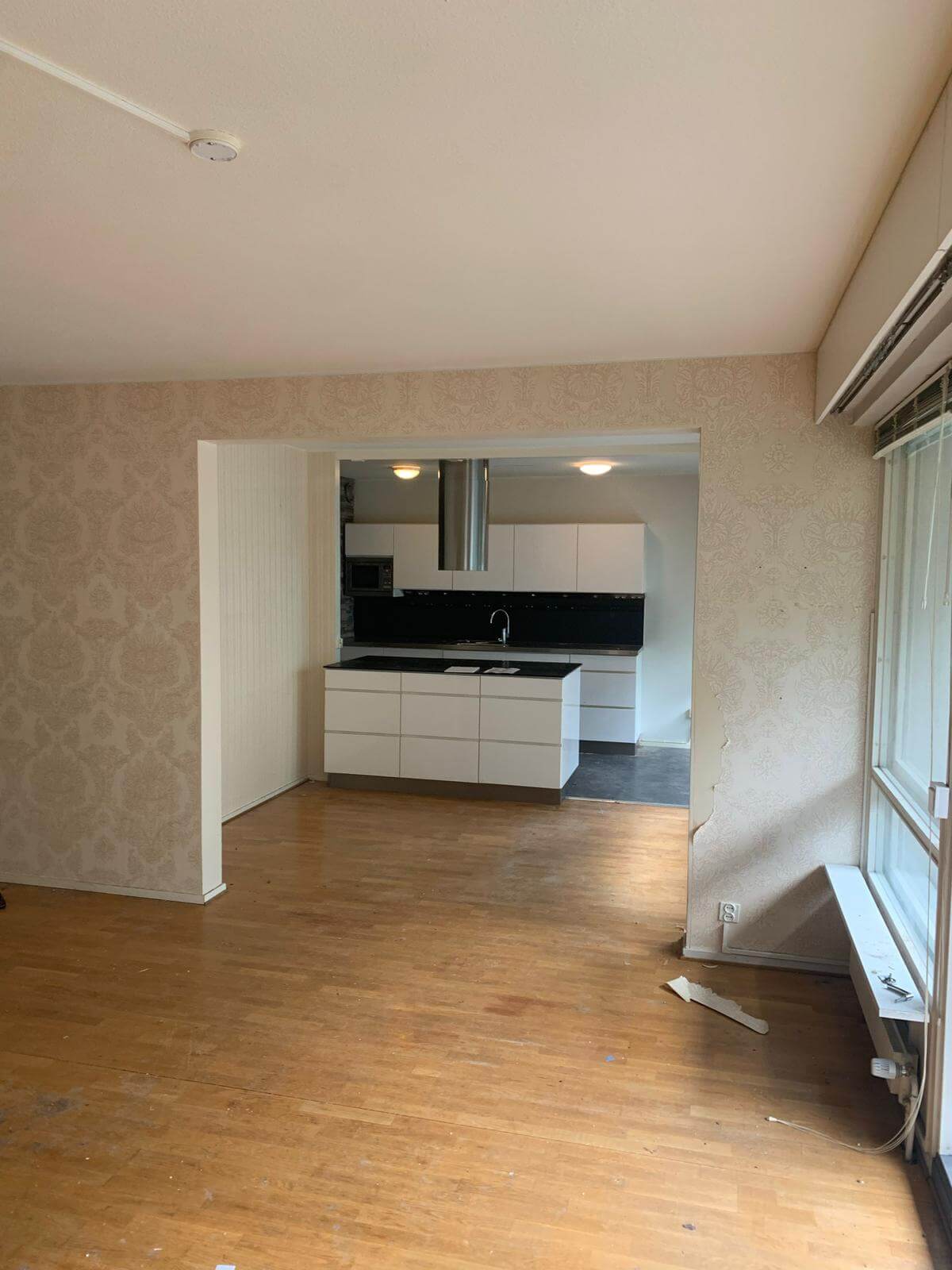 Huone, jossa on puulattia, tapetoidut seinät ja avoin keittiö, jossa on valkoiset kaapit ja saareke.