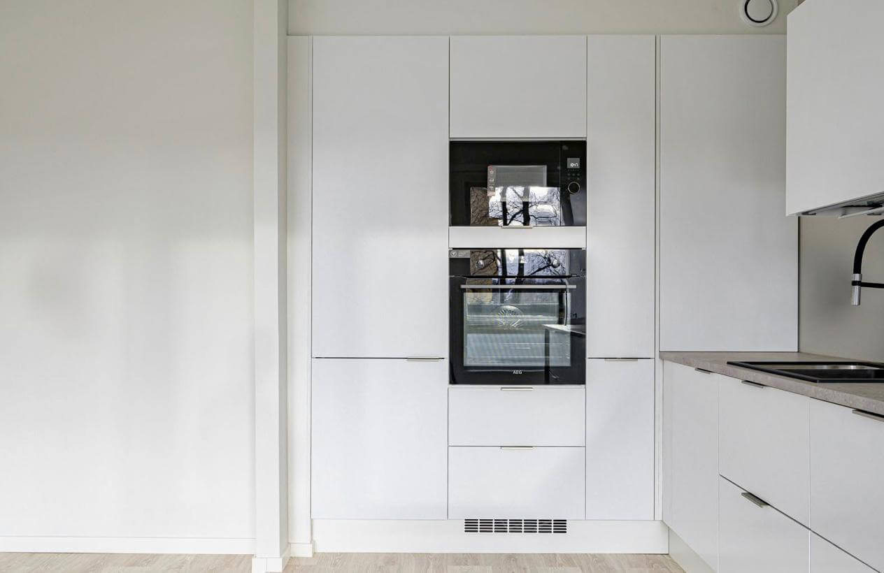 Valkoiset keittiön kaapit, uuni ja mikroaaltouuni, vaalea puulattia ja valkoinen seinä.