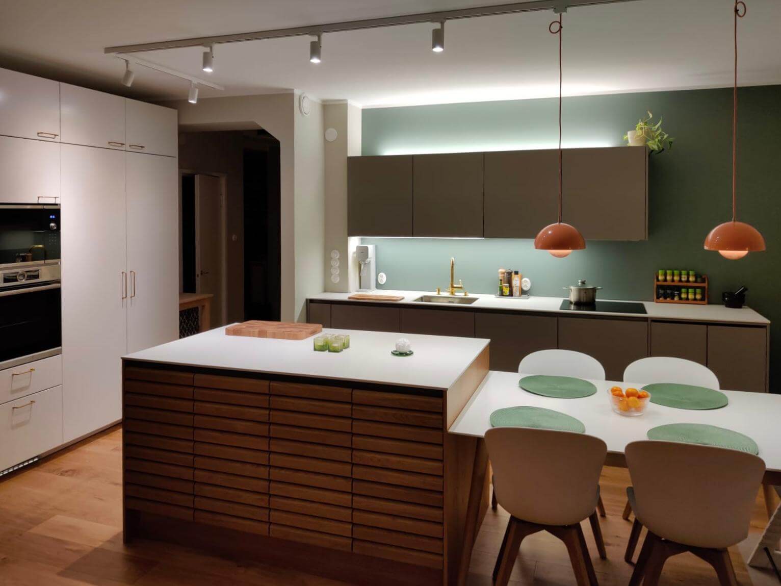 Moderni keittiö, jossa on valkoiset kaapit, puinen saareke, ruskeat yläkaapit, vihreät seinät ja oranssit riippuvalaisimet.