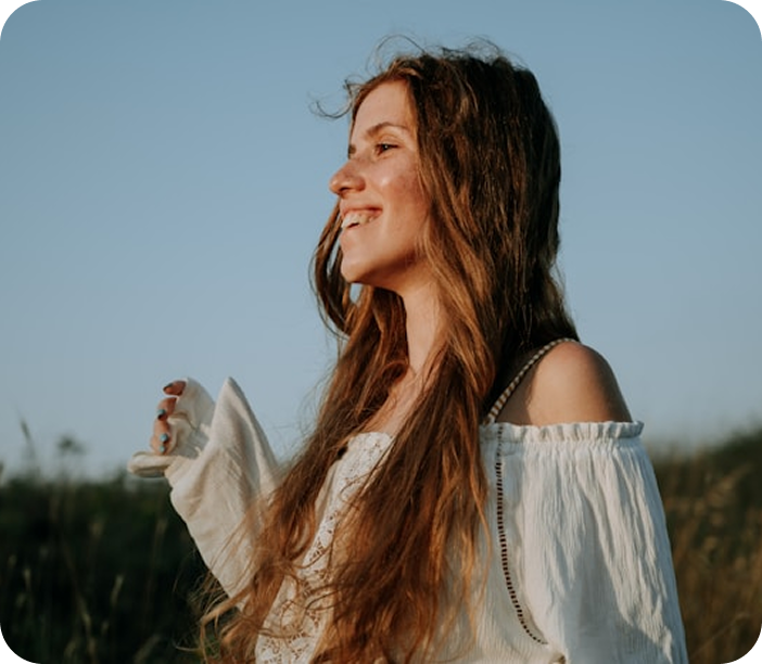 Smiling woman with long brown hair wearing a white off-shoulder top outdoors at sunset.