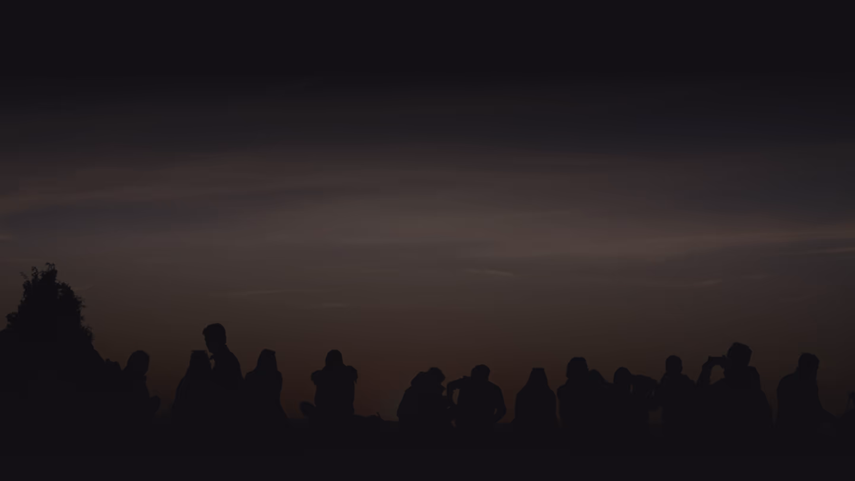 Silhouettes of people sitting and standing outdoors against a darkening sky at dusk.