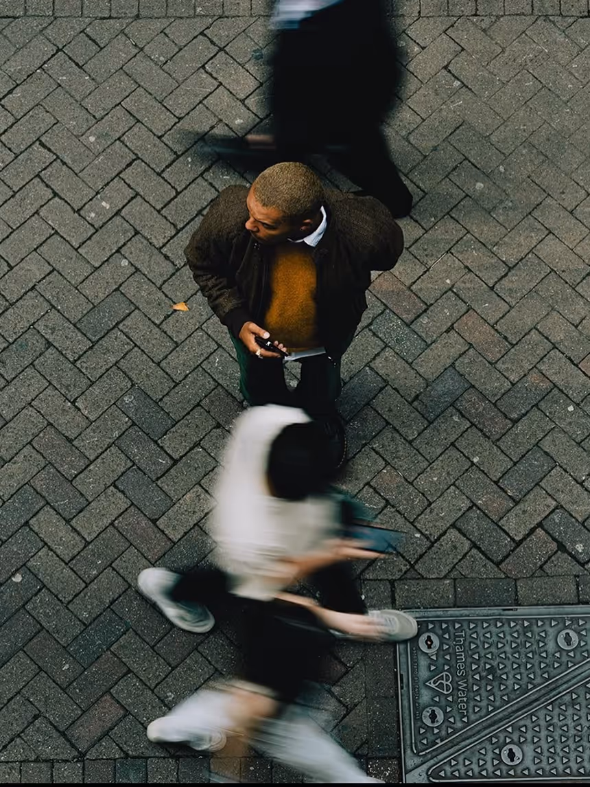 Overhead view of a man standing on a brick sidewalk looking to his left while holding a smartphone, with blurred pedestrians walking past him.