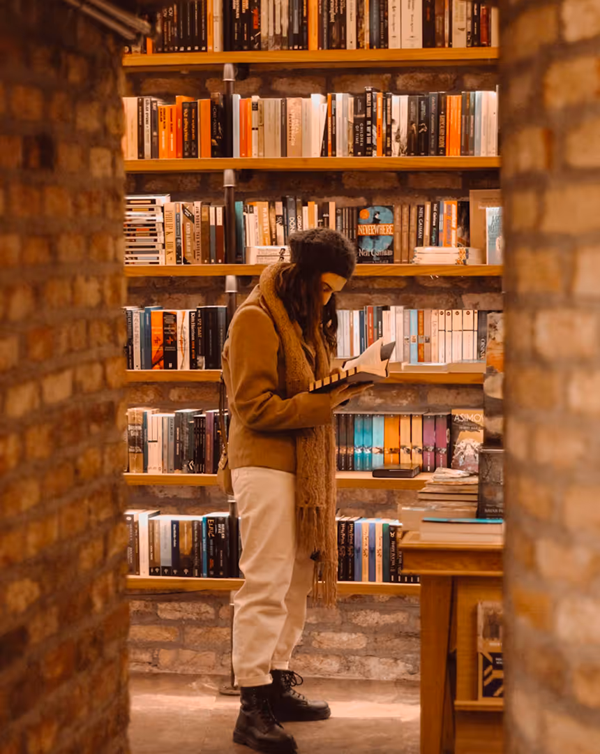 Person wearing a brown jacket, beige pants, and a black beanie standing between brick walls, reading a book in a warm-lit bookstore with wooden shelves filled with books.