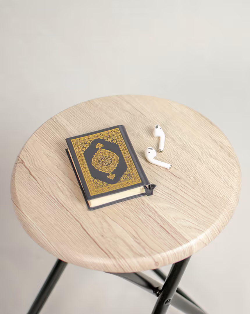 Wooden round table with a closed Quran book featuring ornate gold designs and a pair of white wireless earbuds placed next to it.