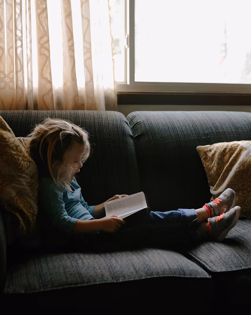 Young child with light hair sitting on a dark couch near a window, reading a book with their legs stretched out and wearing sneakers with pink shoelaces.