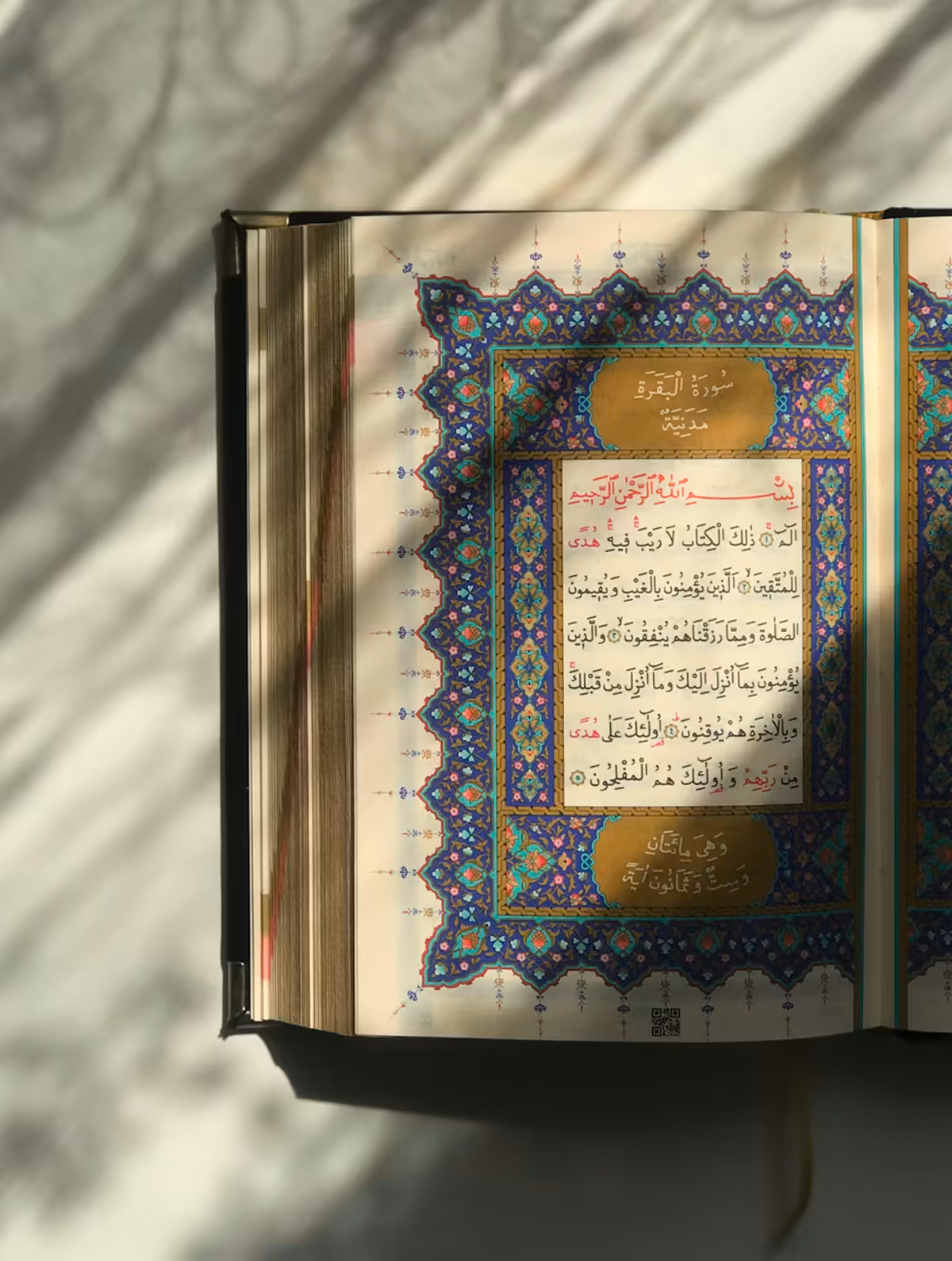 Open Quran showing an ornate, illuminated page with Arabic calligraphy, featuring Quranic text surrounded by an intricate blue, purple, and gold geometric border, with decorative golden oval cartouches containing chapter information.