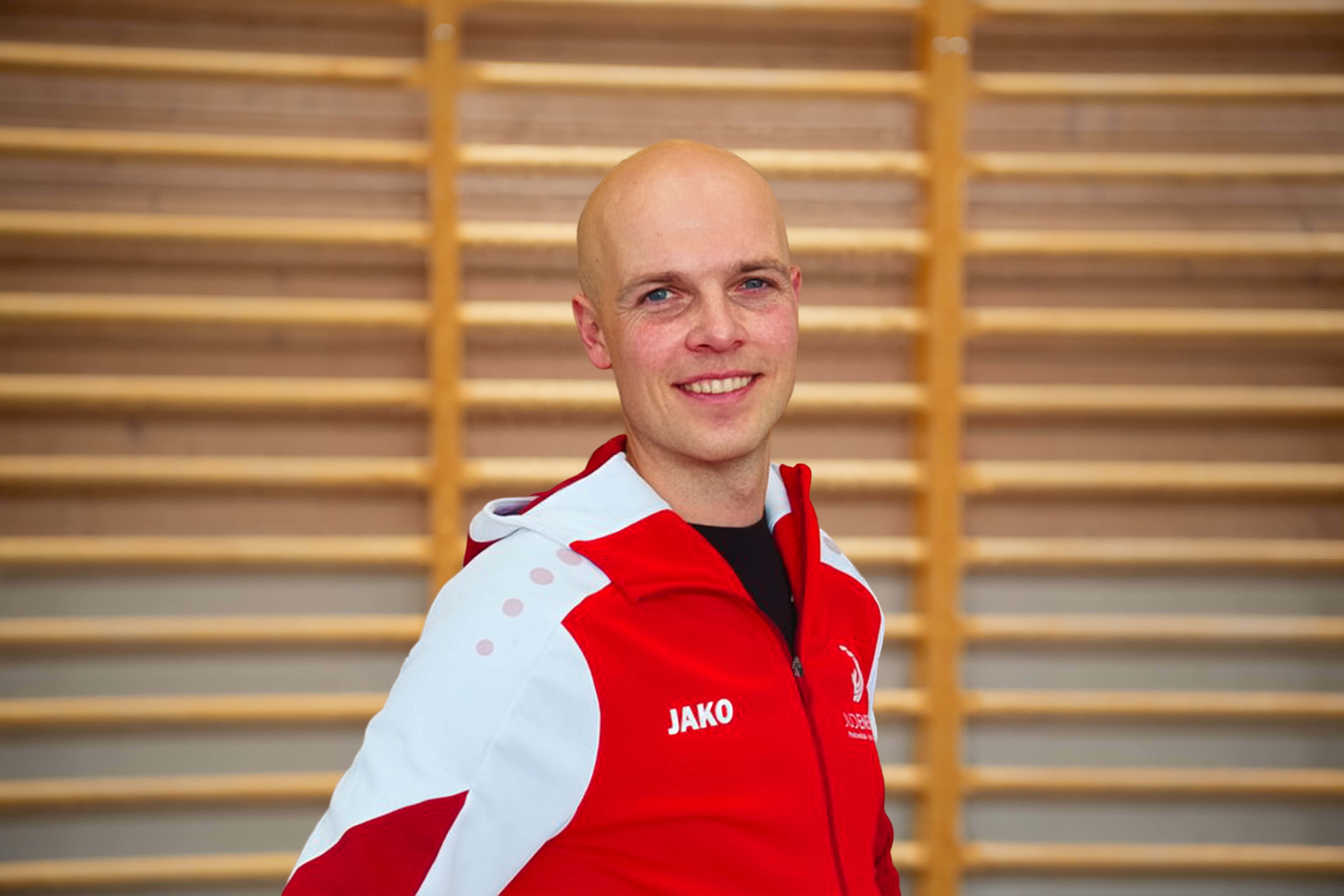 Ein lächelnder Mann mit Glatze trägt eine rote und weiße Trainingsjacke vor einer Holzstangenwand.