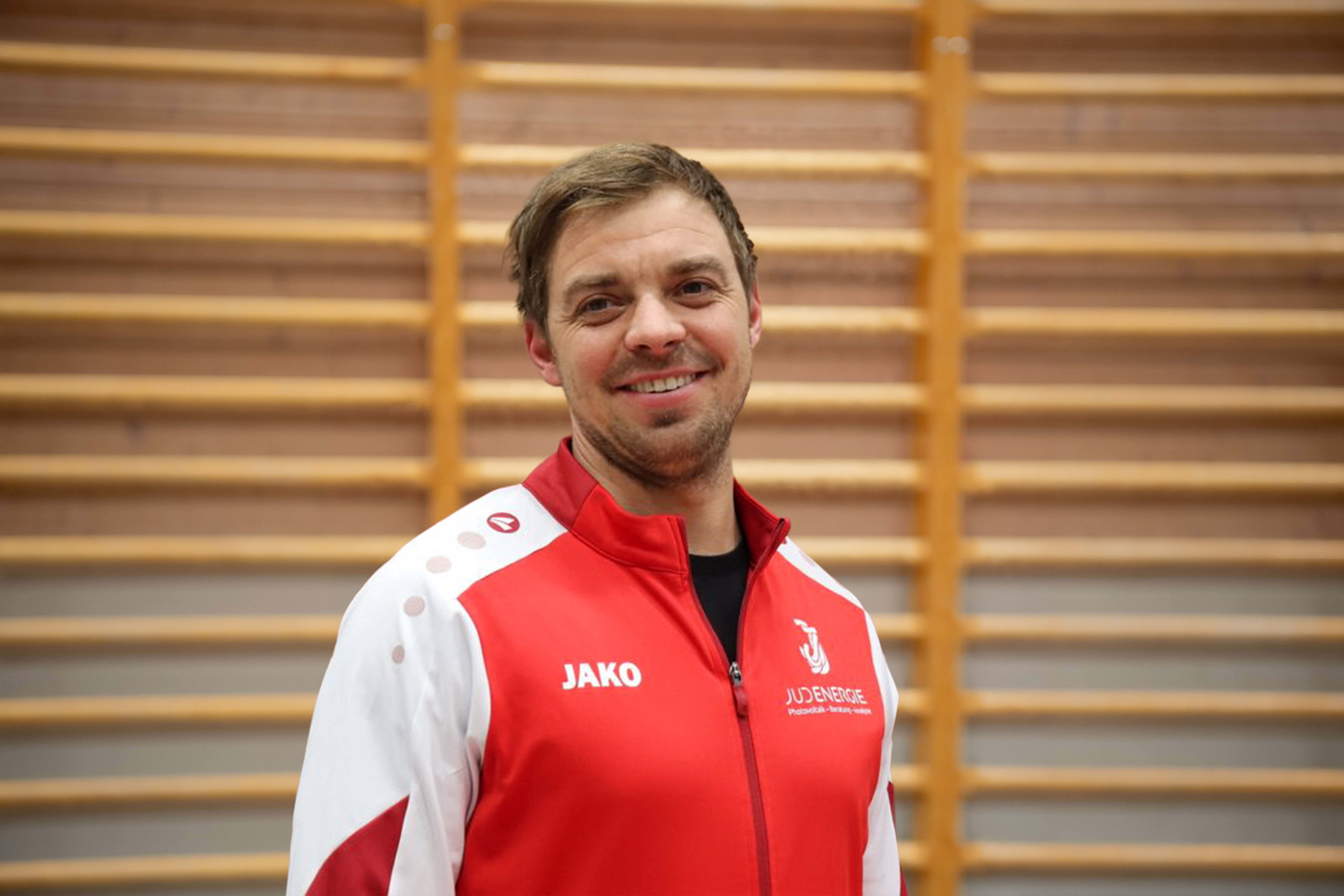 Mann lächelt in roter und weißer Sportjacke vor einer Holzwand mit Sprossen.