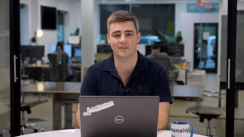 Man sitting at a desk using a Dell laptop with a 'NoVenture' sticker, in an office with other people working in the background.