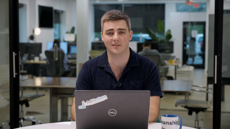 Man sitting at a desk using a Dell laptop with a 'NoVenture' sticker, in an office with other people working in the background.