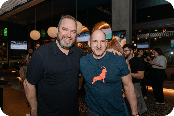 Two smiling men standing close together in a lively indoor setting with people and TV screens in the background.