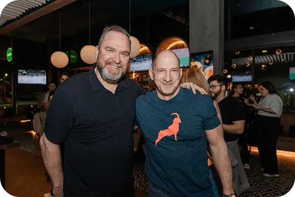 Two smiling men standing close together in a lively indoor setting with people and TV screens in the background.