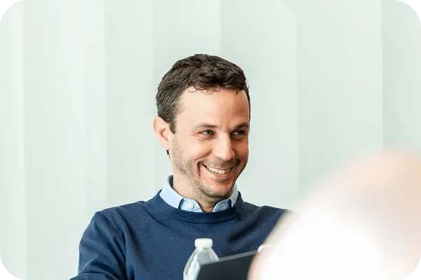 Smiling man with short dark hair wearing a blue sweater over a collared shirt, sitting indoors with a water bottle and tablet in front of him.