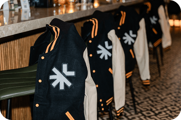 Row of black and beige varsity jackets with orange accents hanging over green bar stools in a modern interior.