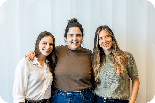 Three smiling women standing closely with arms around each other against a light patterned wall.