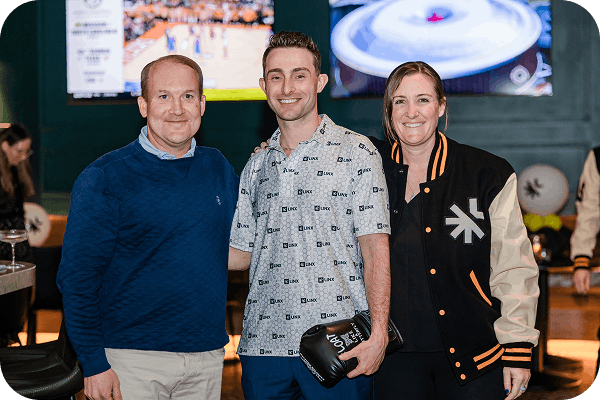Three people standing and smiling indoors with a sports game on TV in the background; the man in the middle is holding a black boxing glove.