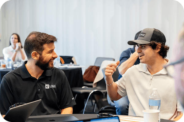Two men smiling and talking across a table in a casual meeting setting, one wearing a black LINX polo and the other a white shirt with a LINX cap.