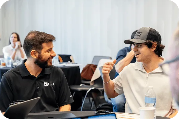 Two men smiling and talking across a table in a casual meeting setting, one wearing a black LINX polo and the other a white shirt with a LINX cap.