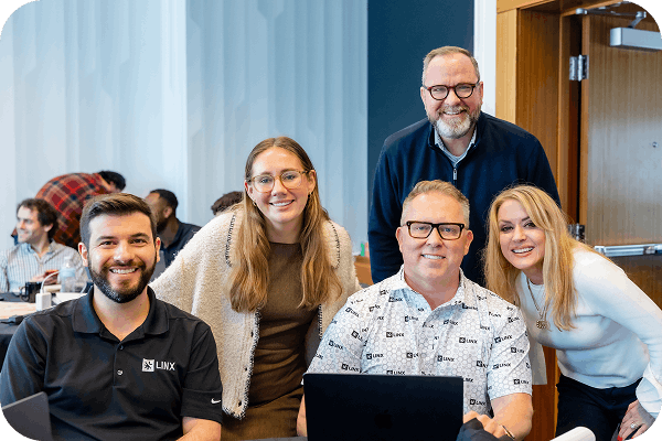 Five smiling colleagues posing together in an office, with a laptop on the table and others working in the background.