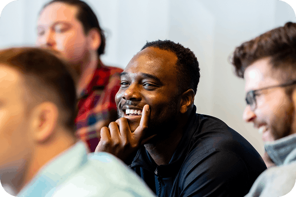 Smiling man with dark skin and short hair resting his chin on his hand while sitting with others.