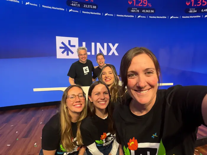 A group of six people smiling and posing for a selfie in front of a blue wall with the "LINX" logo at the Nasdaq MarketSite. They're wearing black shirts with colorful designs, conveying a joyful and celebratory mood.