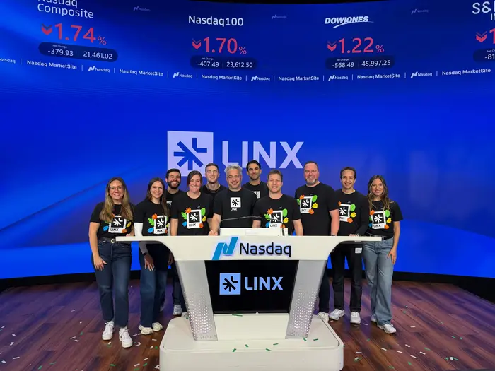 A group of ten smiling people stand behind a Nasdaq podium, wearing matching shirts. The backdrop displays falling stock indices, creating a celebratory yet serious mood.