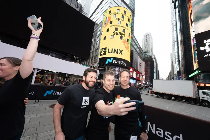 Three people smiling and taking a selfie in Times Square, New York, with a Nasdaq billboard displaying "LINX" and "$83M" in the background. The mood is celebratory.