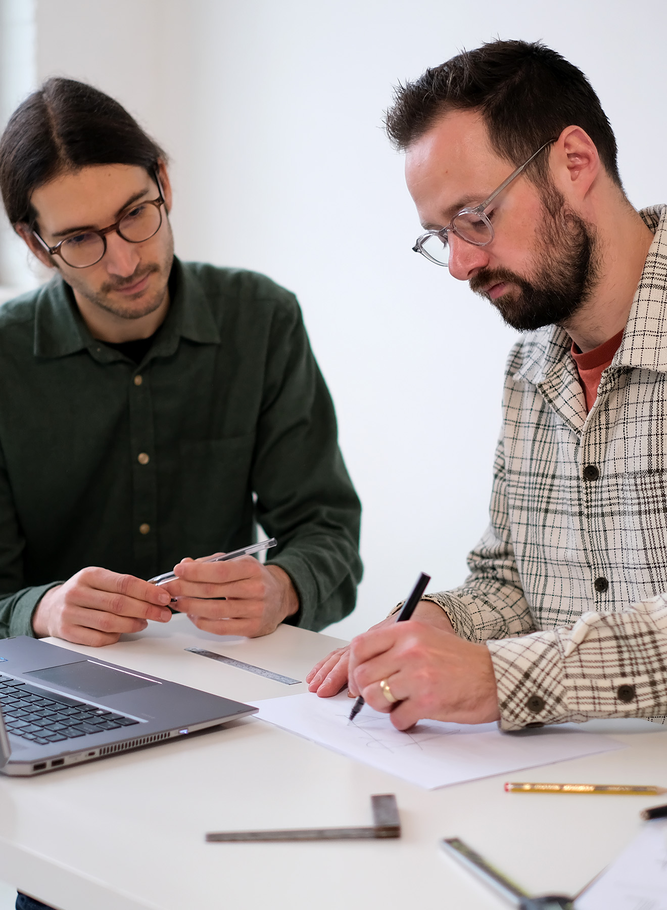 Two men wearing glasses work together at a table with a laptop and drafting tools, one drawing on paper and the other holding a pen.