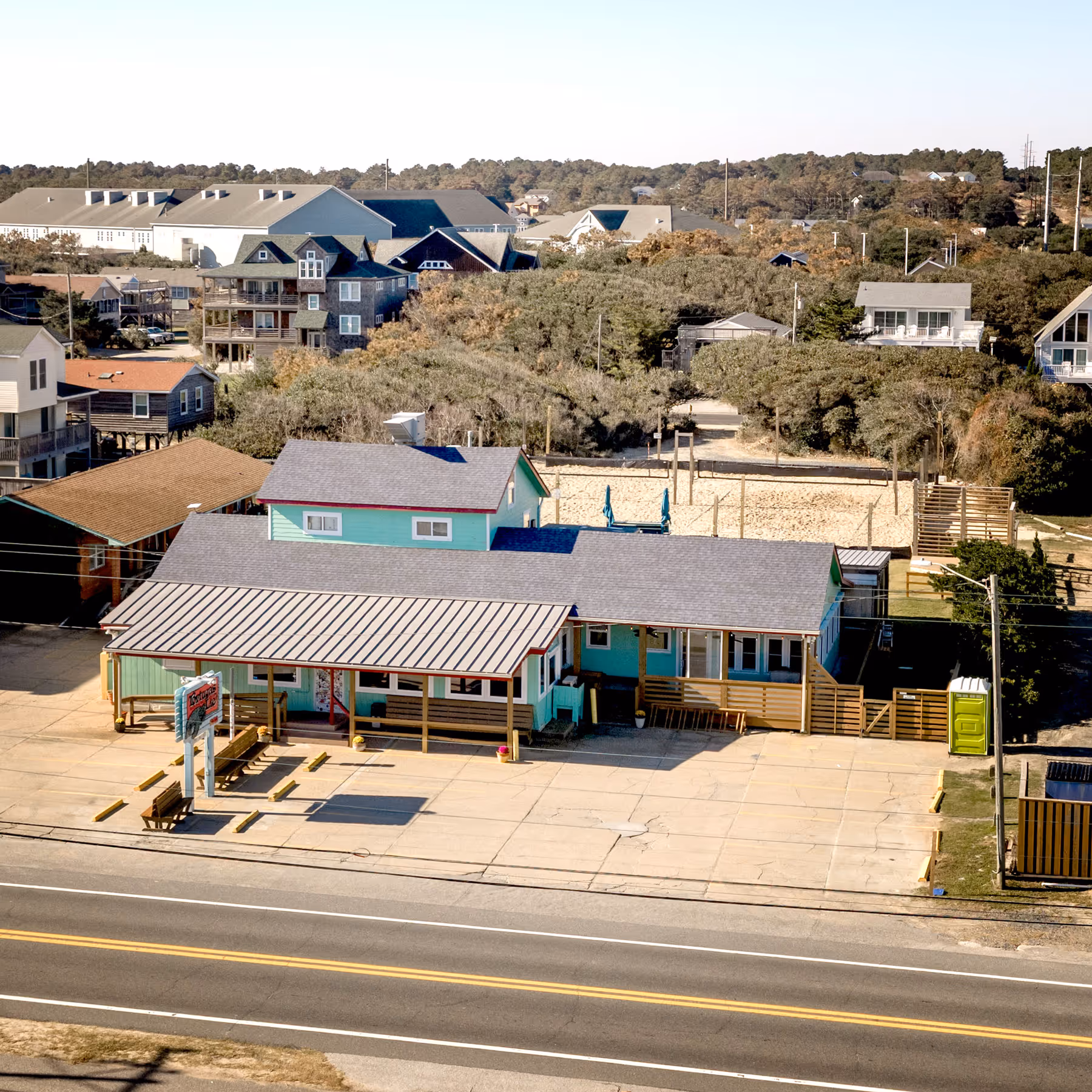 Aerial view of a turquoise building with a metal roof and empty parking lot beside a two-lane road in a residential area.