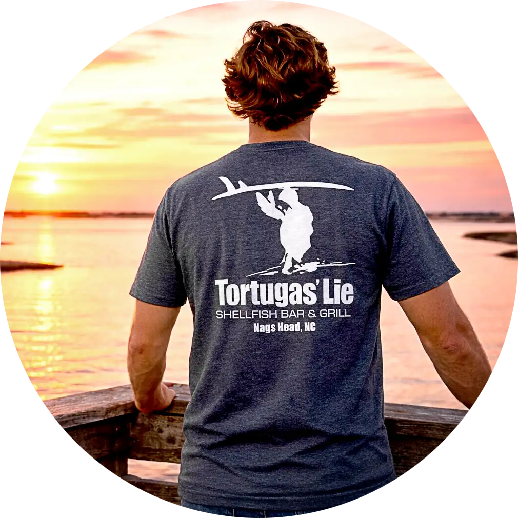 Person with short wavy hair wearing a dark shirt with Tortugas' Lie Shellfish Bar & Grill logo standing on a wooden railing overlooking a sunset over water.